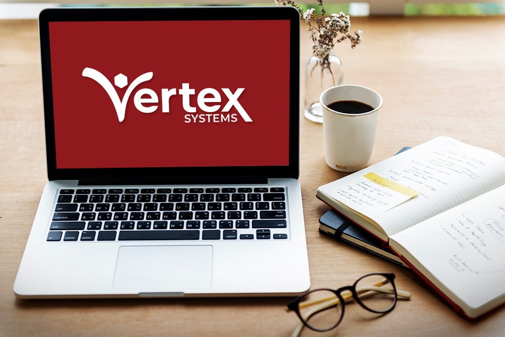 A computer laptop on a desk with glasses and coffee cup. Monitor shows Vertex logo.
