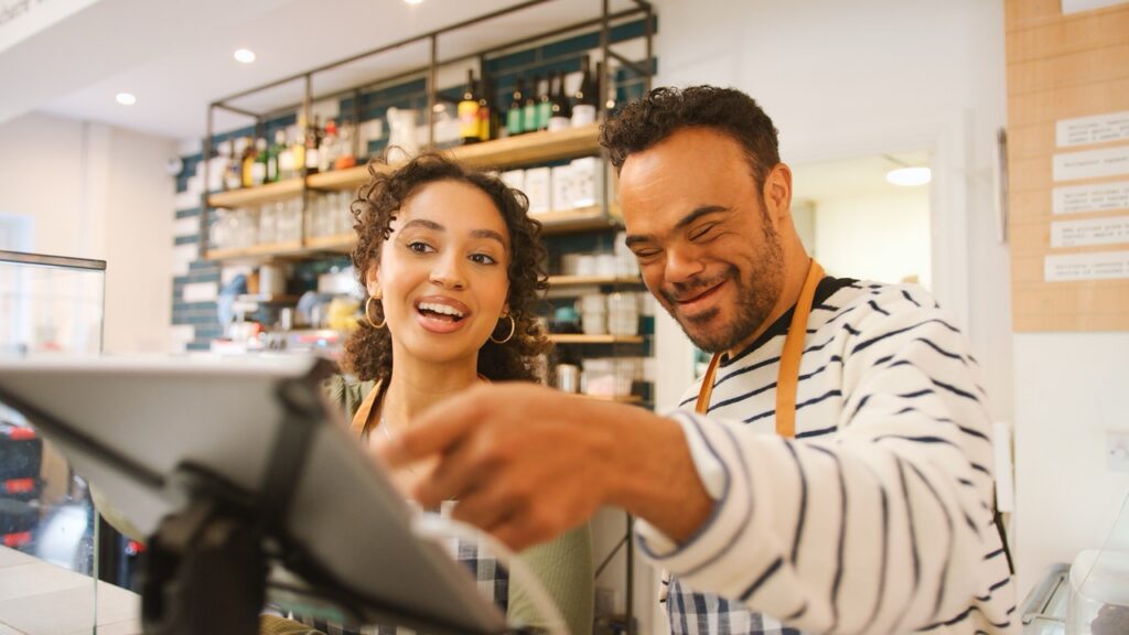 Young woman working a café register alongside a coworker, showing independence for people with IDD through supported employment